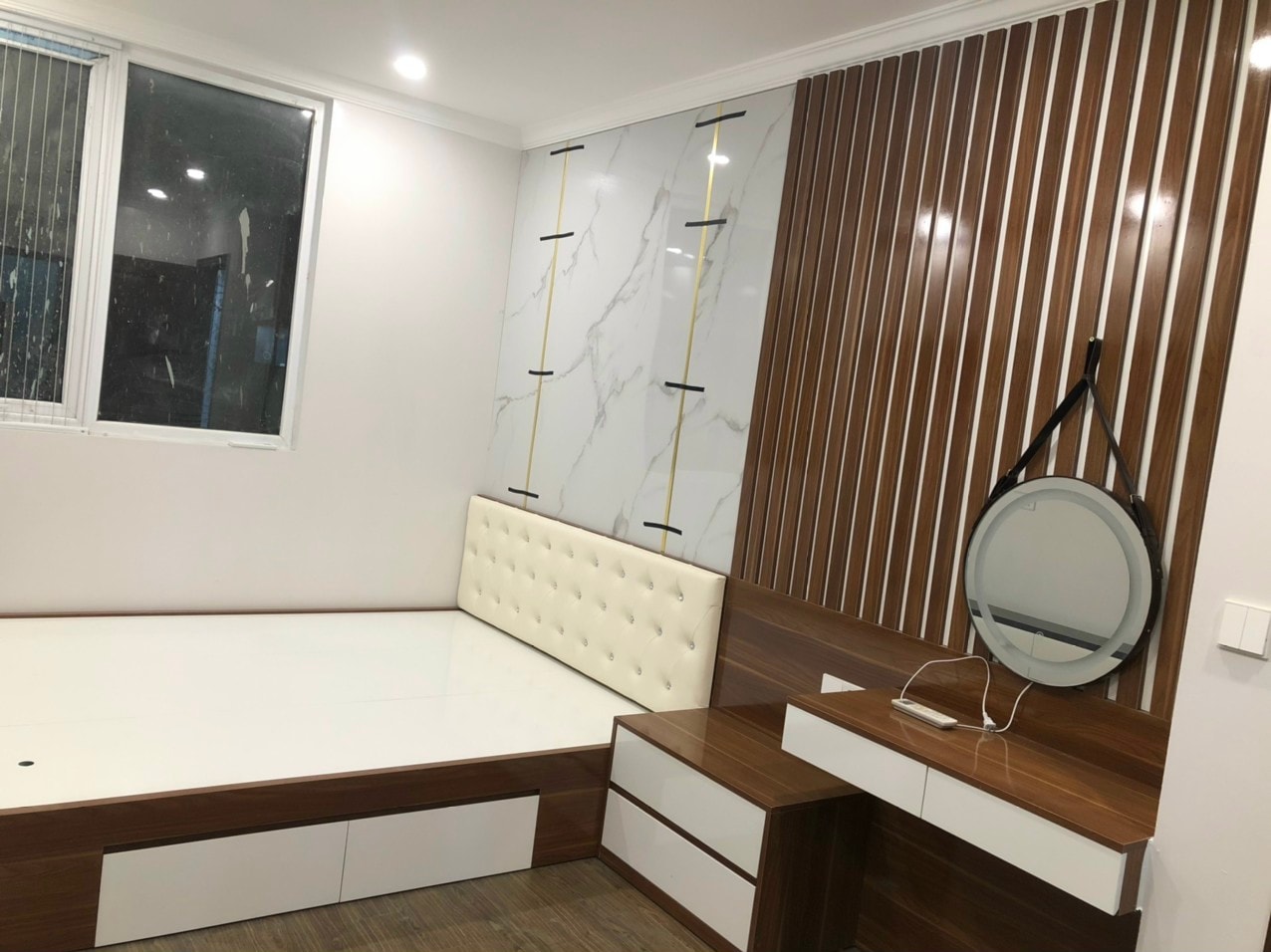 Phòng ngủ gỗ MDF với mảng vách nan sọc dọc, giường ngủ tựa nệm nỉ và gương tròn treo, góc nhìn 2